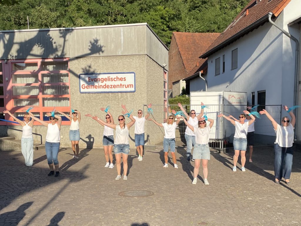 Eine Gruppe in weißen Oberteilen und Jeans tanzt mit blauen Bändern vor dem Evangelischen Gemeindezentrum